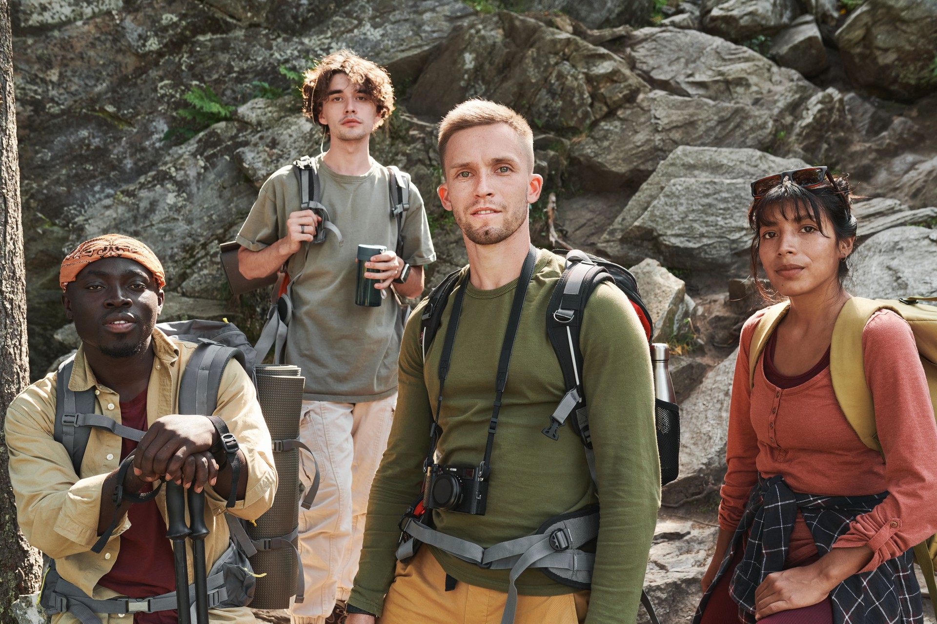 Group of confident young multi-ethnic hikers with bags standing against rocks and looking at camera